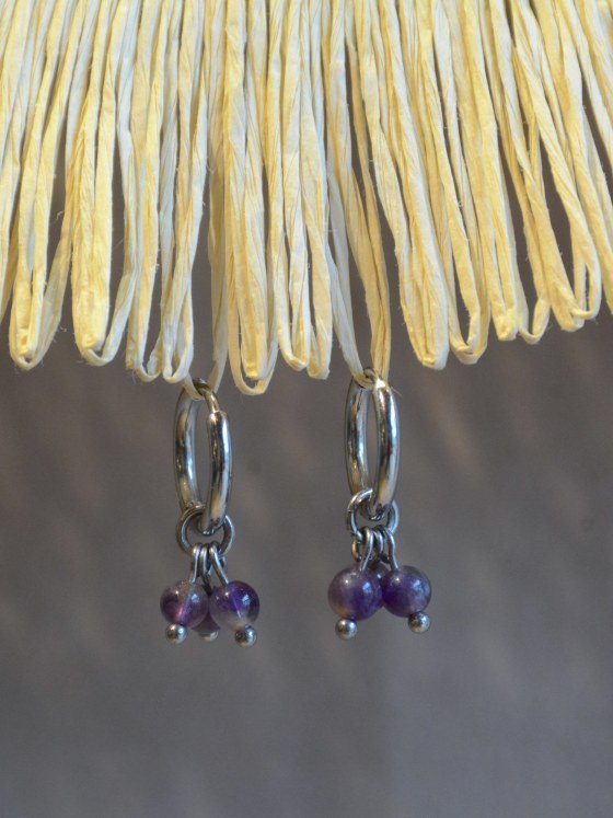 Aretes Belai Amatista Trio, argollas de acero plateado con racimo de tres cuentas redondas de amatista violeta colgando de la argolla, apoyados sobre fibra de rafia y lino
