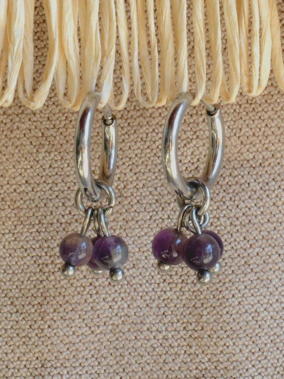 Aretes Belai Amatista Trio, argollas de acero plateado con racimo de tres cuentas redondas de amatista violeta colgando de la argolla, apoyados sobre fibra de rafia y lino