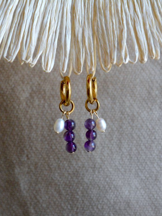 Aretes Belai Amatista y Perla, argollas de acero con racimo de tres cuentas en abanico: amatista violeta, perla blanca y amatista violeta, apoyados sobre fibra de rafia y lino