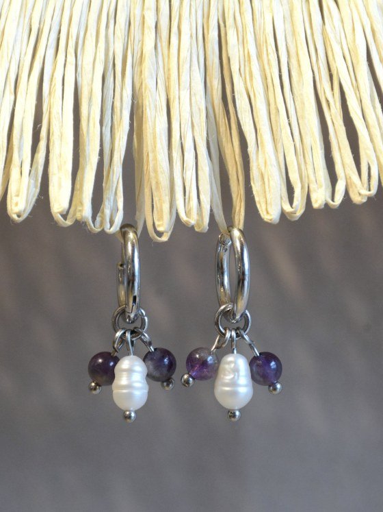 Aretes Belai Amatista y Perla, argollas de acero plateado con racimo de cuentas de amatista violeta y una perla blanca en el centro, apoyados sobre fibra de rafia y lino