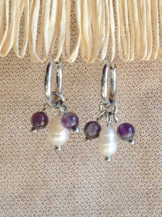 Aretes Belai Amatista y Perla, argollas de acero plateado con racimo de cuentas de amatista violeta y una perla blanca en el centro, apoyados sobre fibra de rafia y lino