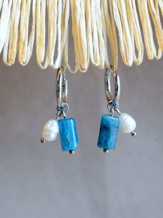 Aretes Belai Apatita y Perla, argollas de acero plateado con dije colgante de piedra apatita azul rectangular y una perla blanca, apoyados sobre fibra de rafia y lino