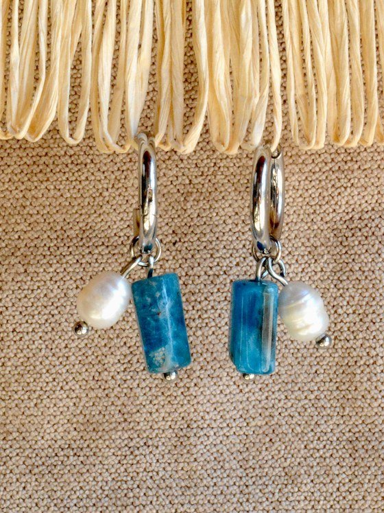 Aretes Belai Apatita y Perla, argollas de acero plateado con dije colgante de piedra apatita azul rectangular y una perla blanca, apoyados sobre fibra de rafia y lino