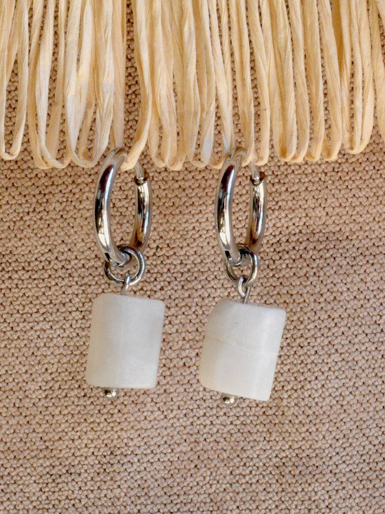 Aretes Belai Piedra Luna, argollas de acero plateado con dije colgante de piedra luna blanca en forma rectangular redondeada y pequeña bolita plateada en el extremo, apoyados sobre fibra de rafia y lino