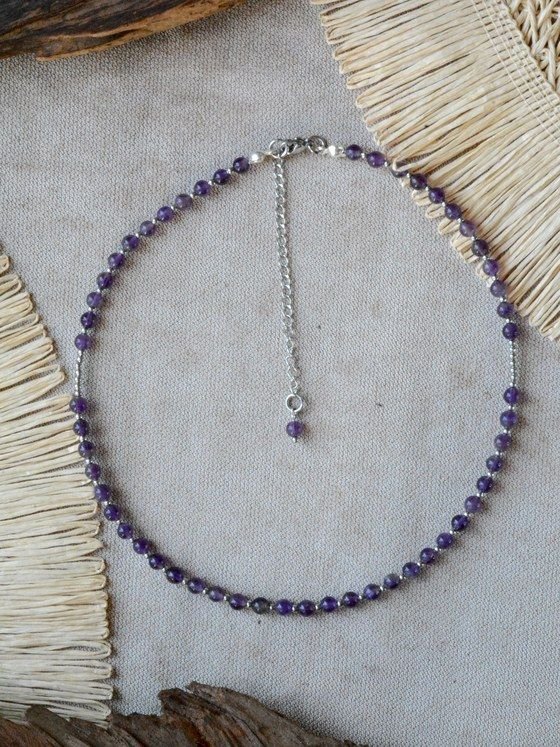 Collar Belai de cuentas redondas de amatista con pequeñas cuentas espaciadoras plateadas, broche de mosquetón plateado y cadena de extensión con una cuenta de amatista colgante, apoyado sobre lino junto a madera y fibra natural