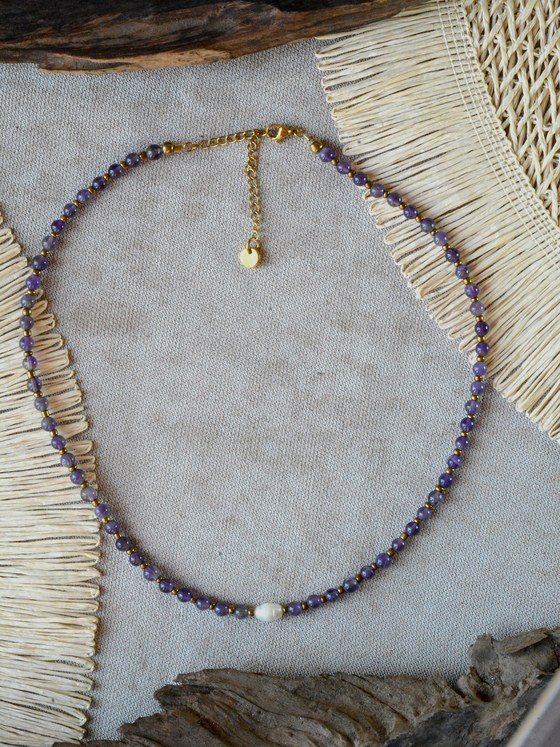 Collar Belai de cuentas redondas de amatista con pequeñas cuentas espaciadoras doradas, una perla blanca como acento asimétrico, broche de mosquetón dorado y cadena de extensión con dije redondo colgante, apoyado sobre lino junto a madera y fibra natural