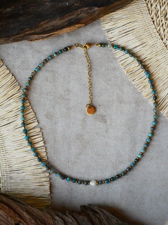 Collar Belai de cuentas redondas de apatita azul con pequeñas cuentas espaciadoras doradas, una perla blanca como acento asimétrico, broche de mosquetón dorado y cadena de extensión con dije redondo colgante, apoyado sobre lino junto a madera y fibra natural
