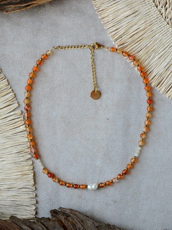Collar Belai de cuentas de cornalina naranja y citrino combinadas con pequeñas cuentas espaciadoras doradas, una perla blanca como acento asimétrico, broche de mosquetón dorado y cadena de extensión con dije redondo colgante, apoyado sobre lino junto a madera y fibra natural