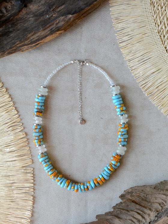 Collar Belai Acra Plateado, cuentas de vidrio reciclado en tono turquesa combinadas con cuentas talladas con motivos florales pintados a mano en naranja y amarillo y vidrio traslúcido esmerilado, broche de mosquetón plateado y cadena de extensión con dije redondo colgante, apoyado sobre lino junto a madera y fibra natural