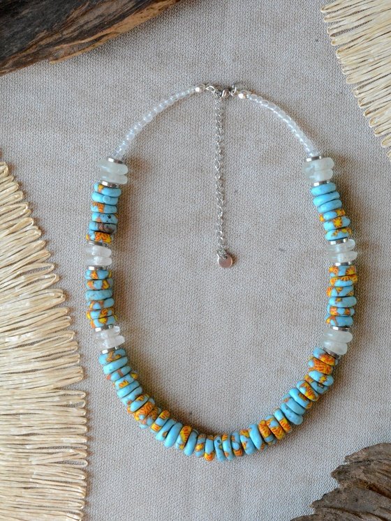 Collar Belai Acra Plateado, cuentas de vidrio reciclado en tono turquesa combinadas con cuentas talladas con motivos florales pintados a mano en naranja y amarillo y vidrio traslúcido esmerilado, broche de mosquetón plateado y cadena de extensión con dije redondo colgante, apoyado sobre lino junto a madera y fibra natural