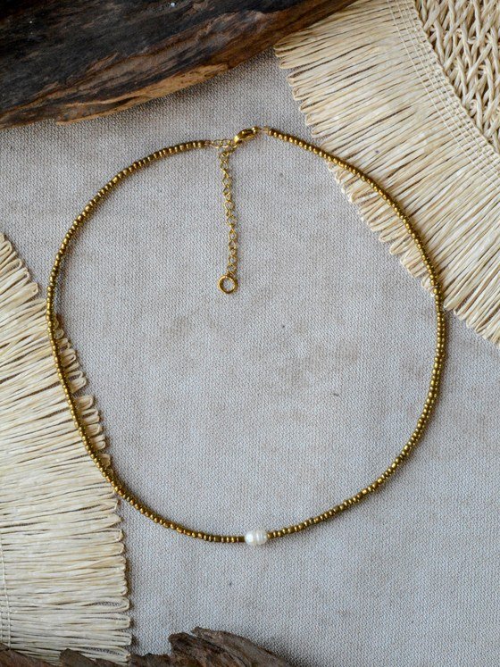 Collar Belai de pequeñas cuentas de vidrio dorado con broche de mosquetón dorado, cadena de extensión con dije redondo colgante y una perla blanca como acento asimétrico, apoyado sobre lino junto a madera y fibra natural