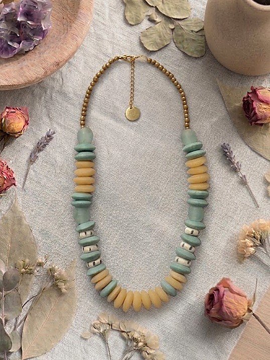 Collar Belai Esmeralda Dorada, cuentas de vidrio reciclado en tono verde salvia con cuentas color mostaza y detalles dorados, apoyado sobre lino junto a flores y hojas secas