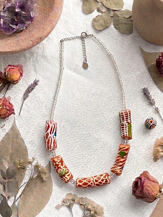 Collar Belai Krobo Rojo, cuentas de vidrio reciclado africano en tono rojo terracota con motivos pintados a mano en blanco y detalles plateados, apoyado sobre lino junto a flores y hojas secas