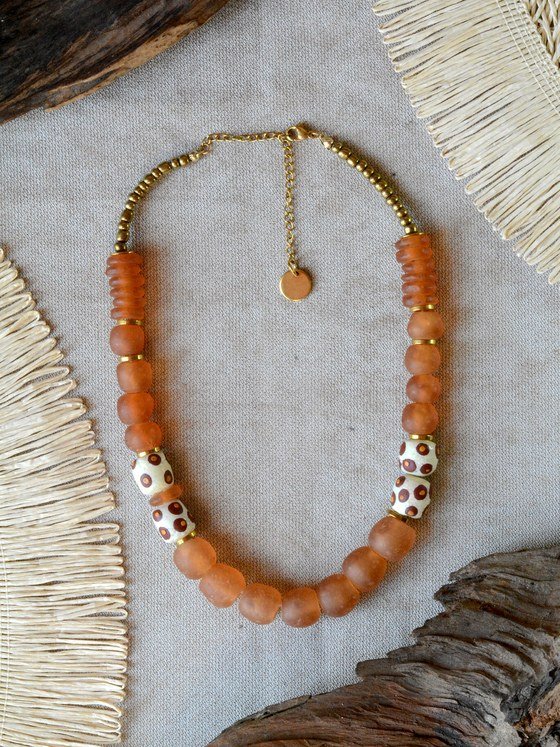 Collar Belai Sohori Dorado, cuentas de vidrio reciclado en tono naranja terracota combinadas con cuentas de hueso pintadas a mano con lunares en tonos marrones, broche de mosquetón dorado y cadena de extensión con dije redondo colgante, apoyado sobre lino junto a madera y fibra natural