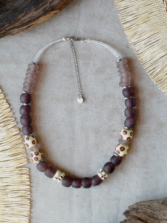 Collar Belai Sohori Violeta, cuentas de vidrio reciclado en tono ciruela violáceo esmerilado combinadas con cuentas de vidrio traslúcido lila y cuentas de hueso pintadas a mano con lunares naranjas, broche de mosquetón plateado y cadena de extensión con dije redondo colgante, apoyado sobre lino junto a madera y fibra natural
