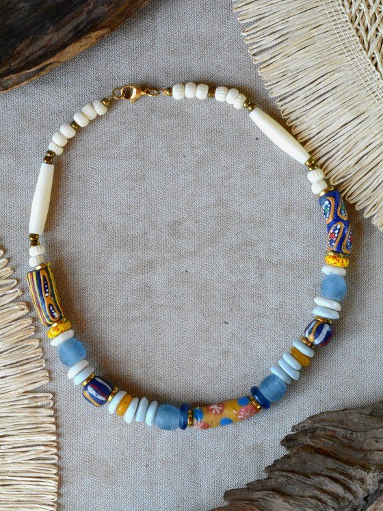 Collar Belai Tere Azul, cuentas de hueso crudo talladas combinadas con cuentas de vidrio reciclado azul aguamarina esmerilado, cuentas Krobo estampadas en azul con motivos florales naranja/amarillo y en amarillo con motivos azul/rojo, separadores dorados, broche de mosquetón dorado y cadena de extensión con dije redondo colgante, apoyado sobre lino junto a madera y fibra natural