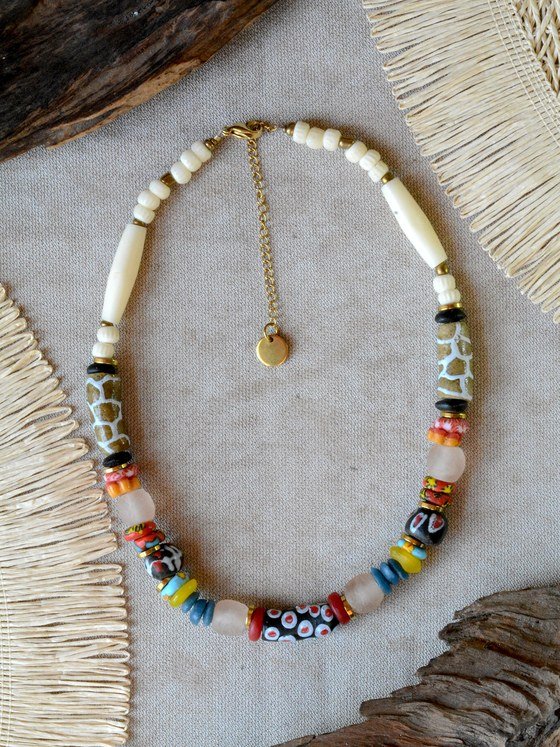 Collar Belai Tere, cuentas de hueso color crudo combinadas con cuentas de vidrio reciclado con motivo tipo jirafa en verde oliva, cuentas estampadas en blanco y negro con lunares rojos, cuentas talladas con flores rojas y amarillas, discos turquesa y amarillos y cuentas de vidrio traslúcido, broche de mosquetón dorado y cadena de extensión con dije redondo colgante, apoyado sobre lino junto a madera y fibra natural