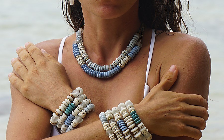 Collar Belai de cuentas de vidrio reciclado en tonos celestes y blancos, puesto sobre el pecho junto a pulseras a juego en la playa
