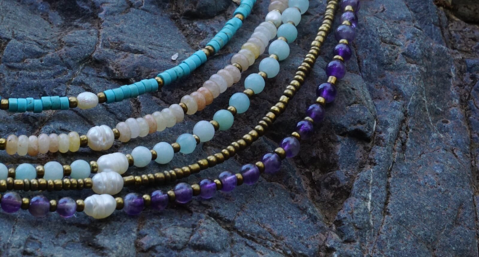 Collares Belai de piedras naturales en tonos turquesa, blanco, amazonita y amatista, con separadores dorados y perlas, apoyados sobre una piedra