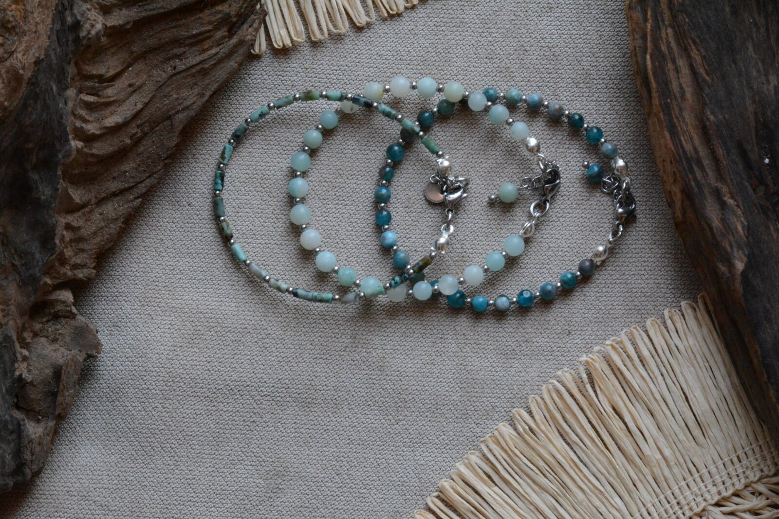 Tres pulseras Belai de piedras naturales en tonos turquesa, verde agua y azul profundo, con cuentas plateadas, apoyadas sobre tela clara junto a madera y fibras naturales