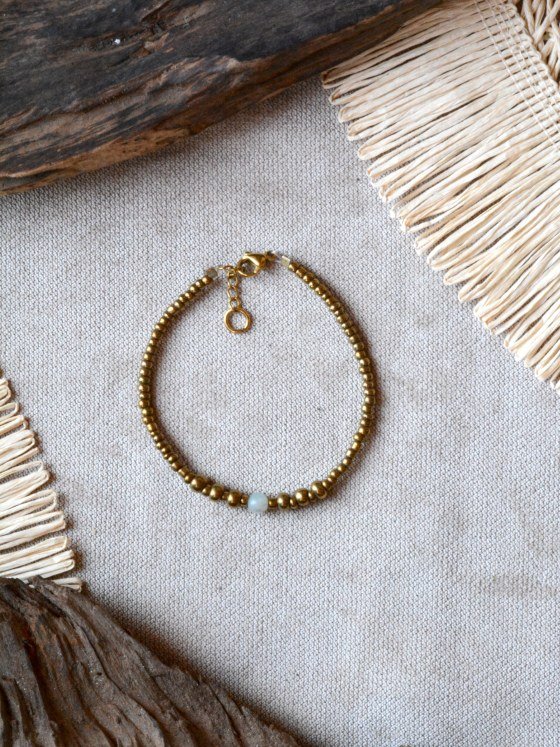 Pulsera Belai Vidrio y Amazonita, cuentas doradas de vidrio con una cuenta de amazonita celeste, apoyada sobre lino junto a madera y fibra natural