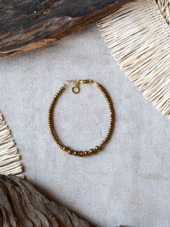 Pulsera Belai Vidrio y Ámbar, cuentas doradas de vidrio con una cuenta ámbar facetada, apoyada sobre lino junto a madera y fibra natural