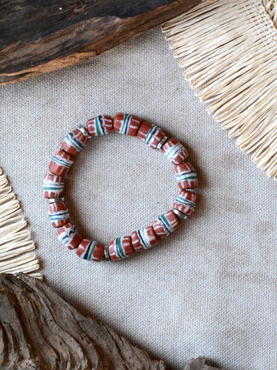 Pulsera Belai Jirapa, cuentas de vidrio reciclado en tono terracota con rayas pintadas a mano en blanco, verde y negro, separadas por anillos plateados, apoyada sobre lino junto a madera y fibra natural