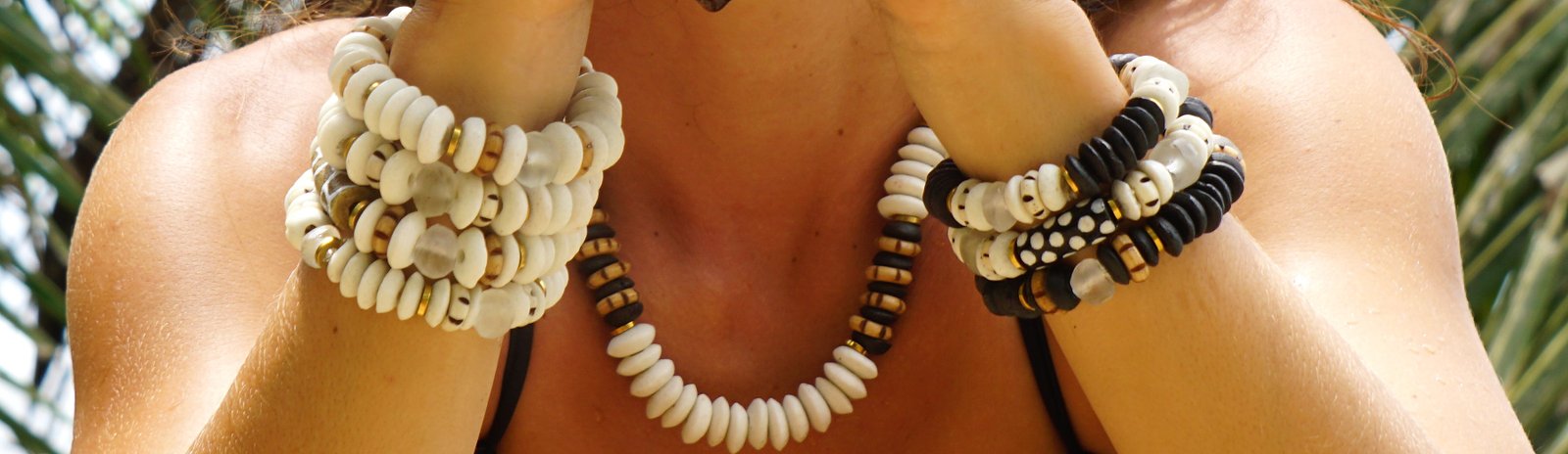 Pulseras Belai de cuentas de vidrio reciclado apiladas en ambas muñecas, junto a un collar de vidrio, sobre fondo de hojas de palmera