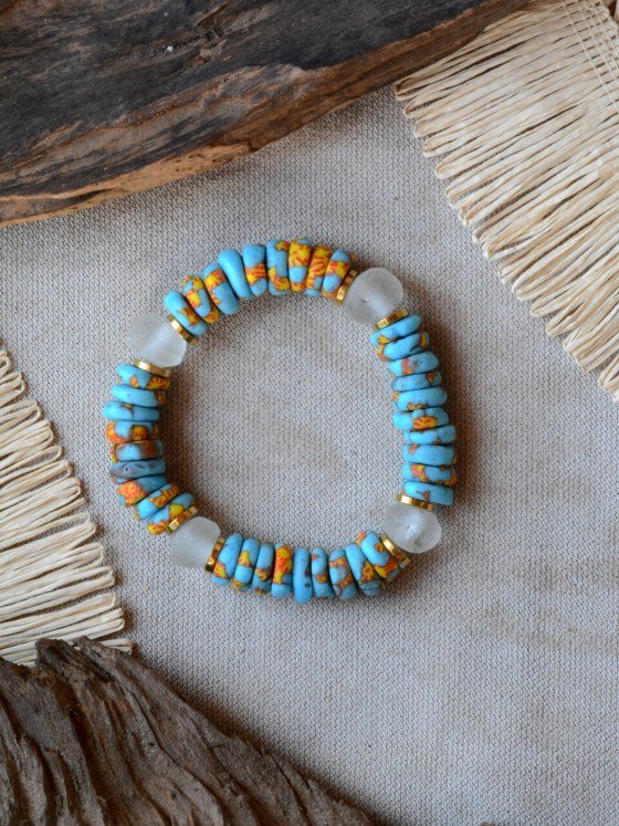 Pulsera Belai Acra, cuentas de vidrio reciclado celeste con motivos pintados a mano en amarillo y naranja, combinadas con cuentas de vidrio traslúcido y anillos dorados, apoyada sobre lino junto a madera y fibra natural