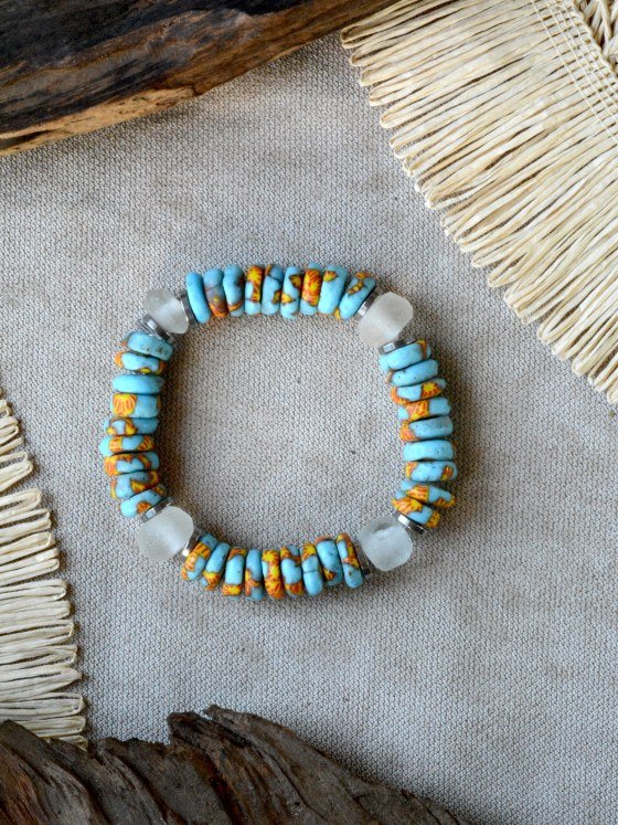 Pulsera Belai Acra Plateada, cuentas celestes de vidrio reciclado con motivos amarillo y naranja pintados a mano combinadas con vidrio traslúcido esmerilado y anillos plateados, apoyada sobre lino junto a madera y fibra natural