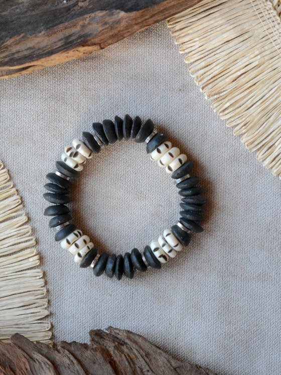 Pulsera Belai Black & White, cuentas de vidrio reciclado negro mate combinadas con cuentas de hueso color crudo con motivos pintados a mano y anillos plateados, apoyada sobre lino junto a madera y fibra natural