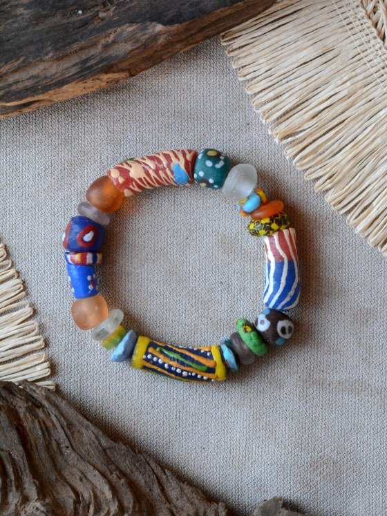 Pulsera Belai Krobo 1, cuentas de vidrio reciclado multicolor pintadas a mano al estilo Krobo, combinando vidrio traslúcido y cuentas pintadas en rojo, azul, verde, amarillo y naranja, apoyada sobre lino junto a madera y fibra natural