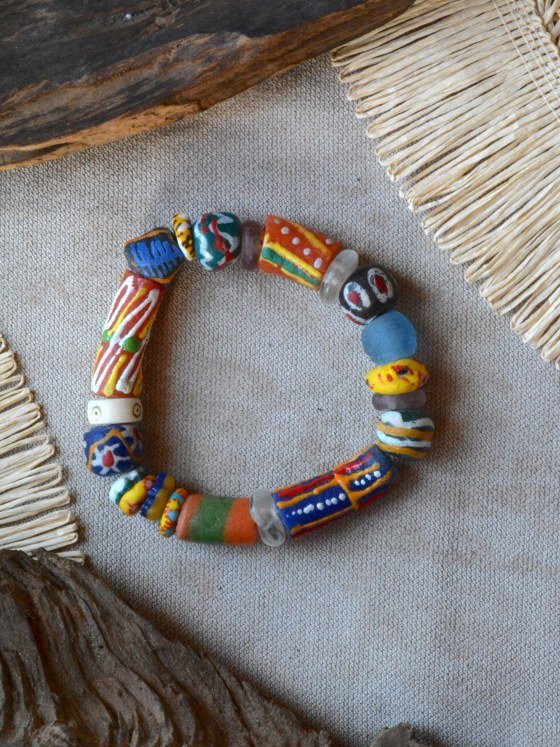 Pulsera Belai Krobo 3, cuentas de vidrio reciclado multicolor pintadas a mano al estilo Krobo, combinando rojo, azul, naranja, amarillo y verde azulado con un anillo de hueso y cuentas de vidrio traslúcido, apoyada sobre lino junto a madera y fibra natural