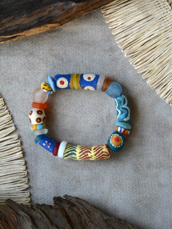 Pulsera Belai Krobo 5, cuentas de vidrio reciclado multicolor pintadas a mano con motivos variados: azul con ojos blanco/rojo y banda dorada envuelta, disco estampado tigre celeste y amarillo, vidrio traslúcido rosa y naranja, blanco con círculos marrón, verde con líneas onduladas, verde con motivo de flor naranja y rojo, rayas rojo/blanco/azul, amarillo con rayas cebra verde oscuro y rojo, apoyada sobre lino junto a madera y fibra natural