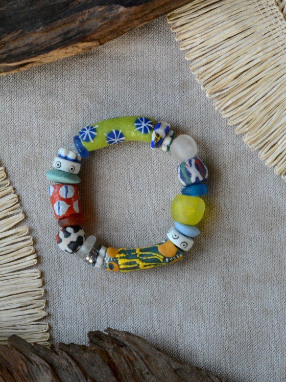 Pulsera Belai Krobo 6, cuentas de vidrio reciclado multicolor pintadas a mano con motivos variados: verde lima con estrellas azules, blanco con ojos y banda azul, verde agua liso, roja con círculos blancos y línea azul, blanco y negro estilo vaca, celeste y vidrio traslúcido, rayas azul/verde/blanco/rojo, celeste, amarillo liso, celeste pálido, blanco con ojos, verde azulado con manchas amarillas y naranjas, anillos plateados, apoyada sobre lino junto a madera y fibra natural