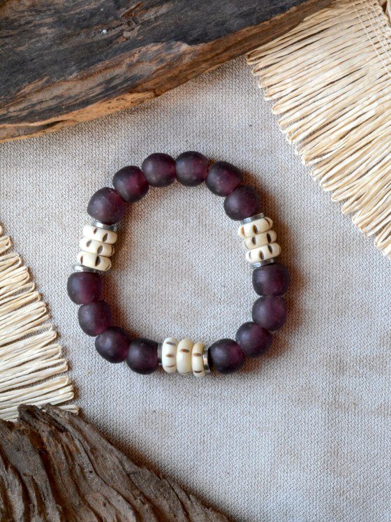 Pulsera Belai Sohori Violeta, cuentas de vidrio reciclado violeta esmerilado combinadas con cuentas talladas en hueso color crudo y anillos plateados, apoyada sobre lino junto a madera y fibra natural