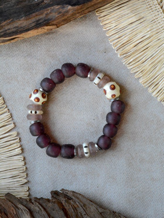 Pulsera Belai Sohori Violeta Krobo, cuentas grandes de vidrio reciclado esmerilado en tono violeta ciruela combinadas con cuentas Krobo color crudo con motivos pintados a mano en marrón y naranja, pequeñas cuentas de vidrio traslúcido rosa lavanda y anillos plateados, apoyada sobre lino junto a madera y fibra natural