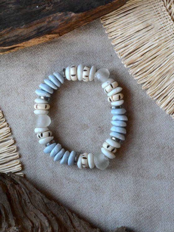 Pulsera Belai Tina, cuentas de hueso color crudo con motivos pintados a mano combinadas con cuentas de vidrio reciclado celeste, vidrio traslúcido y anillos plateados, apoyada sobre lino junto a madera y fibra natural