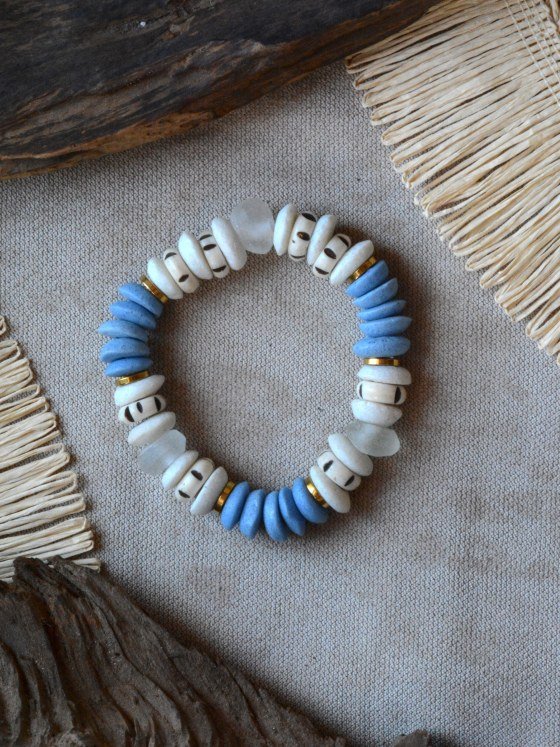 Pulsera Belai Tina Azul, cuentas de hueso color crudo con motivos pintados a mano combinadas con cuentas de vidrio reciclado azul, vidrio tralúscido y anillos dorados, apoyada sobre lino junto a madera y fibra natural