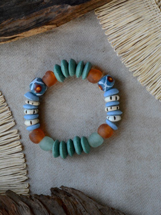 Pulsera Belai Tina Krobo Azul, cuentas de vidrio reciclado verde azulado (discos acanalados) combinadas con cuentas Krobo con motivos pintados a mano en tonos azul y rojo, cuentas de hueso color crudo con marcas talladas, vidrio traslúcido naranja y celeste pálido, apoyada sobre lino junto a madera y fibra natural