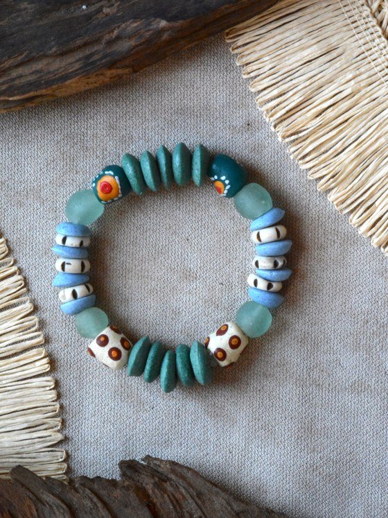 Pulsera Belai Tina Krobo Verde, cuentas de vidrio reciclado verde azulado combinadas con cuentas Krobo pintadas a mano en crudo con motivos naranja y rojo, cuentas celestes con detalle de ojos, y vidrio traslúcido, apoyada sobre lino junto a madera y fibra natural