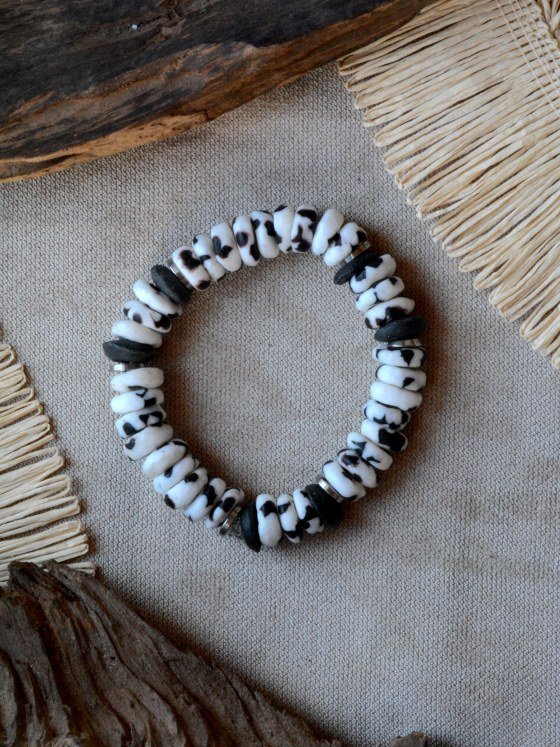 Pulsera Belai Zebra, cuentas blancas con manchas irregulares en negro al estilo animal print, combinadas con cuentas de piedra oscura y anillos plateados, apoyada sobre lino junto a madera y fibra natural
