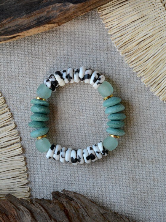 Pulsera Belai Zebra Esmeralda, cuentas blancas con motivos negros estilo animal print combinadas con cuentas de vidrio reciclado verde esmeralda, vidrio traslúcido celeste y anillos dorados, apoyada sobre lino junto a madera y fibra natural
