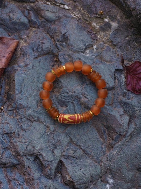 Pulsera Belai Sohori Krobo N, cuentas de vidrio reciclado en tono ámbar con una cuenta central pintada a mano en rojo y dorado, apoyada sobre una piedra con hojas secas